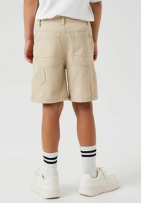 Shorts beiges avec deux poches à l'arrière, une couture simple et une coupe droite, associés à des chaussettes rayées blanches et des baskets blanches.