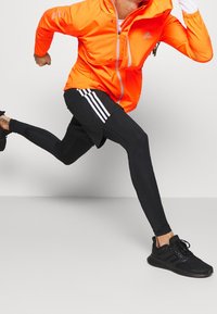 Giacca arancione con zip e dettagli riflettenti, leggings neri con tre strisce bianche e scarpe da corsa nere.