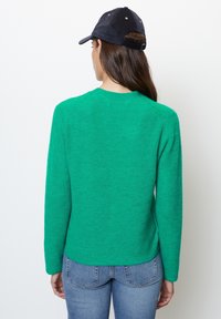 Suéter de punto verde con mangas largas, textura acanalada, cuello redondo y dobladillo ligeramente curvado. Llevado con una gorra azul marino y vaqueros.