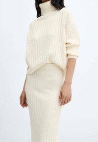 Pull à col roulé en maille côtelée de couleur crème associé à une jupe midi assortie, tous deux en tissu doux et texturé avec une coupe décontractée.