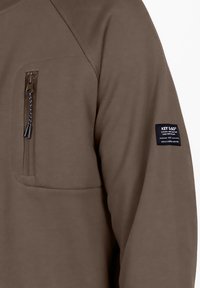 Sudadera de felpa marrón con un bolsillo en el pecho con cremallera, con tejido texturizado y un parche con el logo en la manga. Diseño clásico de cuello redondo.