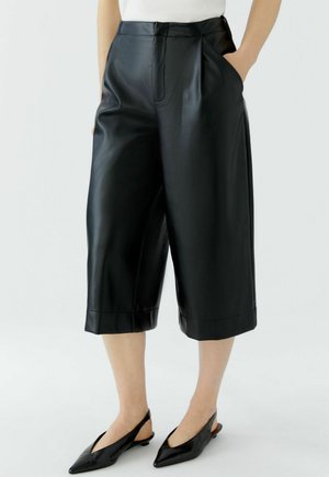 Personne portant un pantalon court en cuir noir à jambes larges et des chaussures slingback noires à bout pointu, debout avec une main dans la poche.