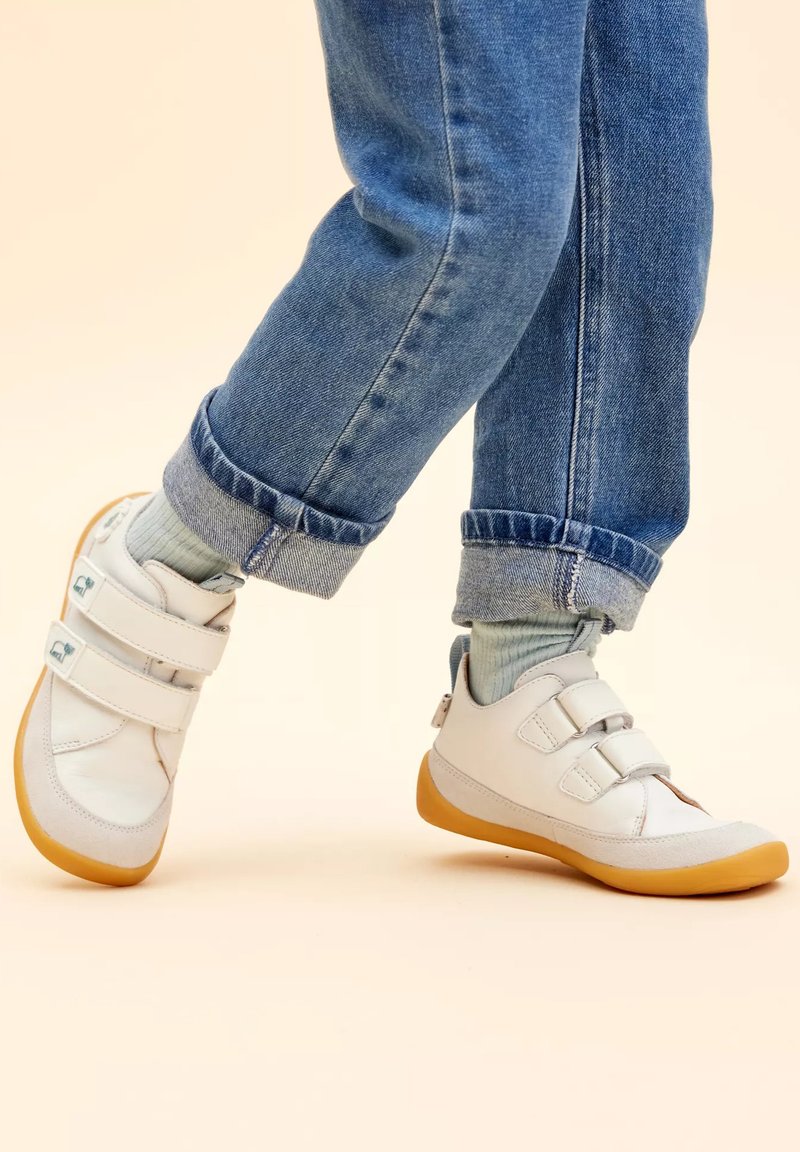 Scarpe bianche in pelle con due fascette a strappo, suola in gomma a contrasto, design arrotondato, abbinate a jeans arrotolati e calze azzurre chiare.
