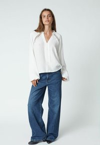Bluză albă cu mânecă lungă, cu un model texturat și închidere cu nasturi. Asortată cu blugi de denim cu croială largă, de nuanță medie albastră, și pantofi negri.
