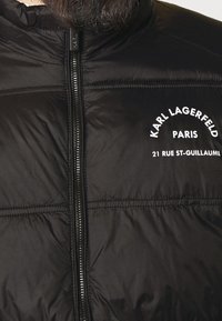 Svart vadderad jacka med hög krage, med dragkedja fram och vit textlogotyp som detaljerar: "KARL LAGERFELD PARIS 21 RUE ST-GUILLAUME."