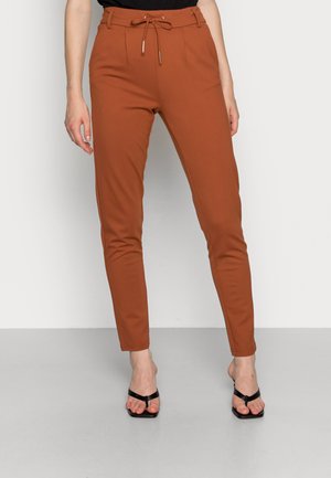Pantalons ajustés marron avec une taille à cordon de serrage et des accents en métal doré. Associés à des sandales noires à lanières. Texture lisse, design élégant.