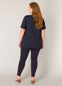 Marineblauwe top met korte mouwen gecombineerd met aansluitende marineblauwe leggings. Model heeft golvend rood haar en draagt beige hakken. Soepele stofstructuur.