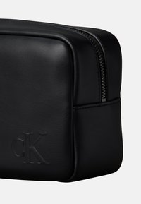 Zwarte leren rechthoekige tas met ritssluiting, voorzien van geperste initialen "cK" en een gladde textuur met subtiele stikseldetails.