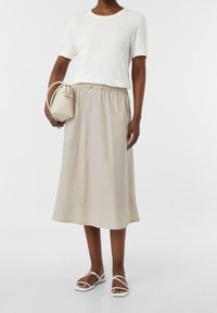 Mujer vestida con una blusa blanca de manga corta, una falda midi beige, sandalias blancas con tiras y sosteniendo un pequeño bolso beige a su lado.