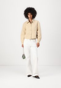 Casaco beige cropped com bolsos frontais e botões grandes, combinado com calças brancas de pernas largas. Pequena mala de mão com detalhes verdes. Sapatos planos pretos.