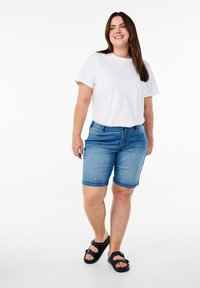 Pantalones cortos de mezclilla azul claro con cinco bolsillos y dobladillos enrollados, combinados con una camiseta blanca lisa y sandalias negras con acentos de tachuelas.