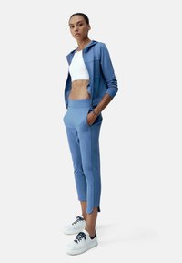 Világoskék, zippzáros kapucnis pulóver zsebekkel, fehér crop top-pal és hozzá illő világoskék, tapered fazonú nadrággal párosítva. Fehér tornacipővel egészül ki az öltözet.