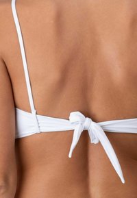 Top bikini bianco con spalline sottili e chiusura posteriore a fiocco. La texture del tessuto è liscia e il design semplice mette in risalto la schiena.
