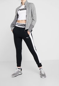 Grå zip-up idrottsjacka över en vit kort topp, parat med svarta byxor med vita ränder och en svart logotyp, samt silvriga sneakers.