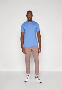 Maglietta blu a maniche corte con una texture mélange, abbinata a pantaloni marrone chiaro a gamba stretta. Scarpe da ginnastica bianche con dettagli neri.