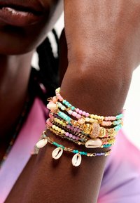 Colorful beaded bracelets in pastel shades with gold accents and shell details. Worn on a brown arm, showcasing a mix of textures and designs.