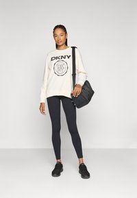 Felpa crema con il logo nero "DKNY", abbinata a leggings neri e sneakers nere. Il modello porta anche una borsa a spalla nera.