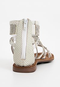 White flat sandal with back zipper, textured snakeskin pattern, rhinestone-embellished ankle strap, and multiple thin straps across foot.