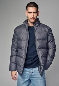 Sivý páperový kabát s vysokým golierom a zipsom, nosený cez tmavý sveter. Model má na sebe modré džínsy a za ním je jednoduché pozadie.
