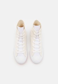 Vysoké tenisky z krémové plátno s bílými tkaničkami, bílou gumovou špičkou a texturovaným textilním svrškem. Ploché bílé švy zvýrazňují design.