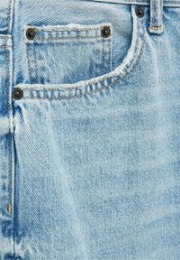 Poche de jean en denim bleu clair avec bords effilochés, coutures visibles et rivets en métal noir aux coins supérieurs.