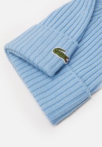 Gorro de malha azul claro com riscas verticais e uma dobra na bainha. Apresenta um logo de crocodilo verde bordado na bainha.