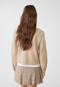 Beige kabát z vlněné směsi s volným střihem a dlouhými rukávy, doplněný hnědou kostkovanou plisovanou sukní s červenými akcenty.