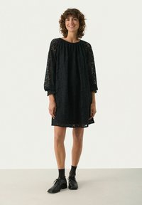 Frau mit lockigem Haar, die ein schwarzes Spitzenkleid mit langen Ärmeln, schwarze transparente Kniestrümpfe und schwarze Schnürschuhe trägt, steht und lächelt.