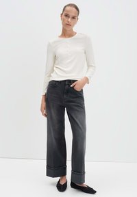 Vrouw gekleed in een witte langekleding henley-shirt, hooggeknoopte zwarte wijde jeans met opgerolde boorden en zwarte instapschoenen, staand tegen een witte muur.