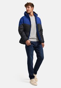 Puffjakke med blå og antracitgrå farveblokke, der har hætte, lynlås lukning og elastiske manchetter. Matchende med grå sweater og mørke jeans.