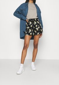 Džínsová košile přes béžové tričko, sladěné s černými květinovými šortkami s žlutými a bílými květy, doplněné bílými teniskami.