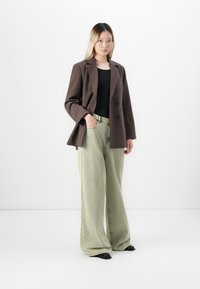 Jeune femme se tient avec une main dans la poche, portant un blazer marron, un top noir, un jean large vert clair et des chaussures noires devant un fond blanc.