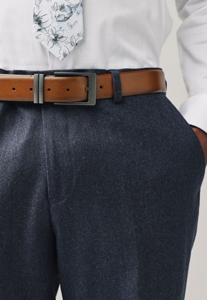 Bruine leren riem met metalen gesp, gedragen over donkerblauwe, gestructureerde wollen broek. Wit overhemd met lange mouwen en een bloemmotief stropdas.