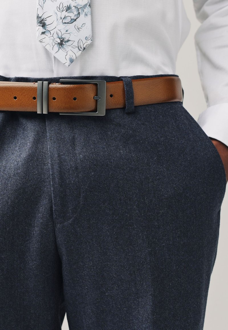 Cintura in pelle marrone con fibbia in metallo, indossata sopra pantaloni di lana blu scuro con texture. Camicia bianca a maniche lunghe con cravatta a motivo floreale.