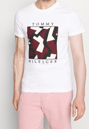 Mann trägt weißes kurzärmeliges T-Shirt mit geometrischem roten, schwarzen und weißen Muster und dem Schriftzug „Tommy Hilfiger“, kombiniert mit hellrosa Hose.