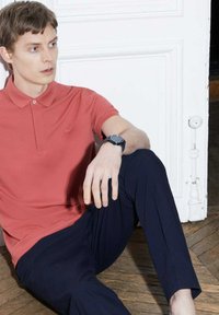 Korallfärgad polo skjorta med krage, liten logotyp på bröstet, tillsammans med marinblå strukturerade byxor. Modell sitter mot en vit vägg.