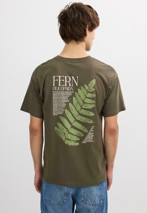 Joven con camiseta verde oliva con un gran gráfico de helecho verde y texto descriptivo en la parte trasera, combinado con vaqueros azul claro.