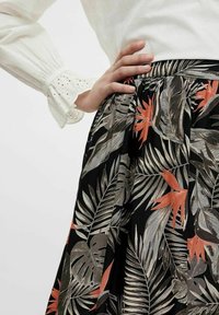 Ruka na bocích, oblečená v bílé blůze s krajkovými manžetami a černé sukni se vzorem tropických listů a oranžových květů.