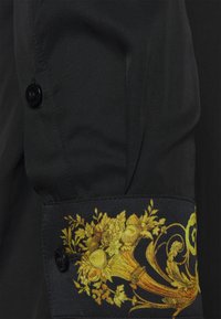 Manga de camisa negra con un botón negro, que presenta un colorido diseño floral en dorado y amarillo en los puños. Tejido texturizado.