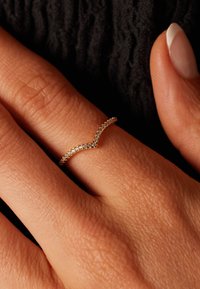 Gouden ring met een golvend ontwerp, bezet met kleine heldere stenen langs de bovenkant. Draagt op een gestructureerde, gebruinde hand tegen een zwarte achtergrond.