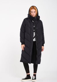 Lange zwarte (puffer) jas met capuchon, ruitvormige stikselpatroon, drukknopen aan de voorkant, zijzakken, gedragen over zwarte leggings en sneakers.