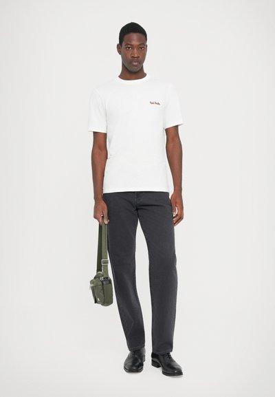 T-shirt bianco in cotone con testo rosso sul petto, abbinato a jeans scuri e scarpe nere, mentre tiene una borsa a tracolla verde. Design minimalista.