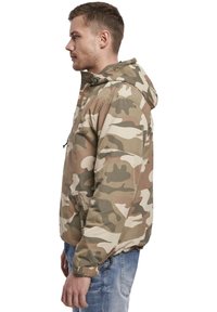 Veste camouflage en beige, vert et tan avec une capuche, des poches zippées et des poignets élastiques. Tissu léger avec une coupe décontractée.