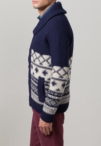 Mörkblå stickad cardigan med sjalkrage, med vita geometriska mönster, en framficka och ribbade ärmslut och nederkant.