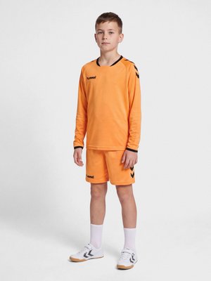 Niño de pie vistiendo una camiseta deportiva naranja de manga larga y pantalones cortos con calcetines blancos y zapatillas deportivas blancas de interior con chevrones negros.