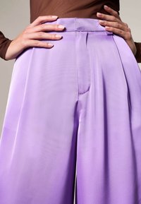 Pantaloni larghi a pieghe in lavanda con finitura satinata liscia. Presentano una vita tailored e tasche laterali nascoste.