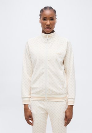 Woman wearing cream-colored zip-up jacket and matching pants with subtle repeating logo pattern, standing against plain white background.