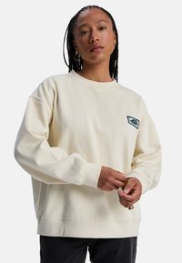 Φούτερ σε κρεμ απόχρωση με απαλό ύφασμα, διαθέτει στρογγυλή λαιμόκοψη και ραφές ώμων που πέφτουν χαμηλά. Μικρό μπλε λογότυπο στο στήθος.