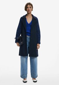 Cappotto navy con maniche in maglia blu testurizzata, abbinato a un top blu a coste e jeans a vita alta azzurri. Gli accessori includono una piccola borsa nera.