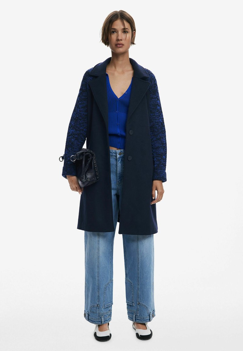 Cappotto navy con maniche in maglia blu testurizzata, abbinato a un top blu a coste e jeans a vita alta azzurri. Gli accessori includono una piccola borsa nera.